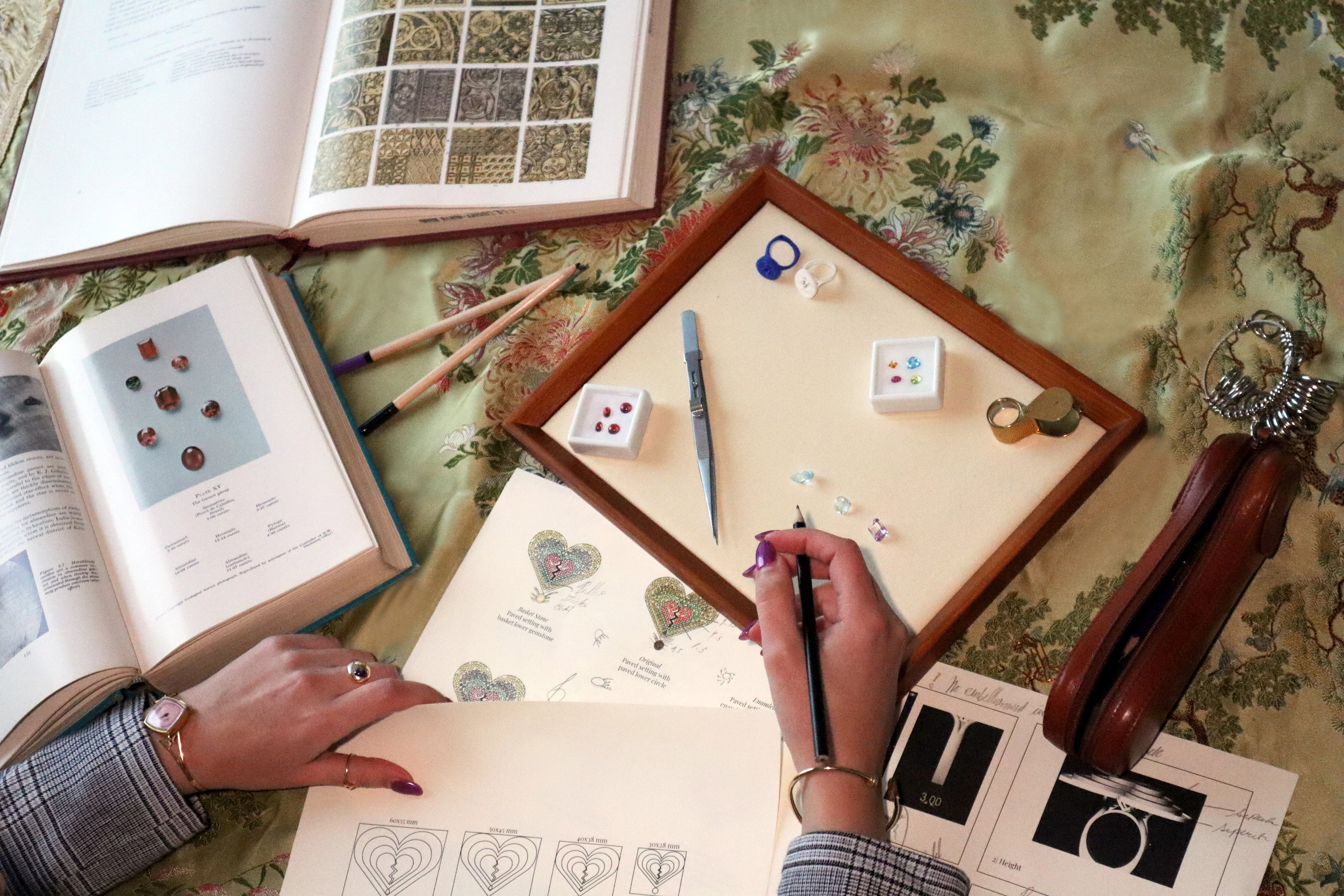 Person working on a craft project with books, tools, and materials on a floral tablecloth.