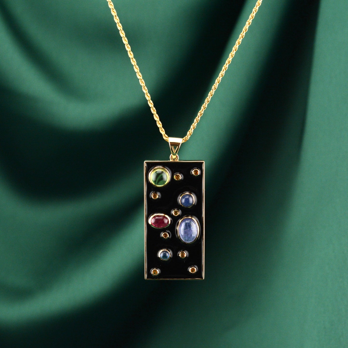 A gold necklace with a rectangular pendant containing multicolored gemstones, displayed against a green draped background.